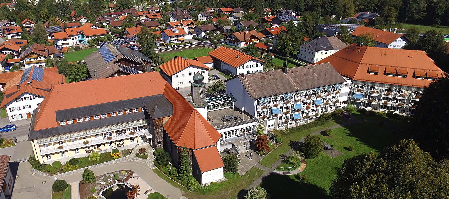 Luftaufnahme eines Gebäudekomplexes mit roten Dächern in einer grünen Landschaft und einem Dorf im Hintergrund
