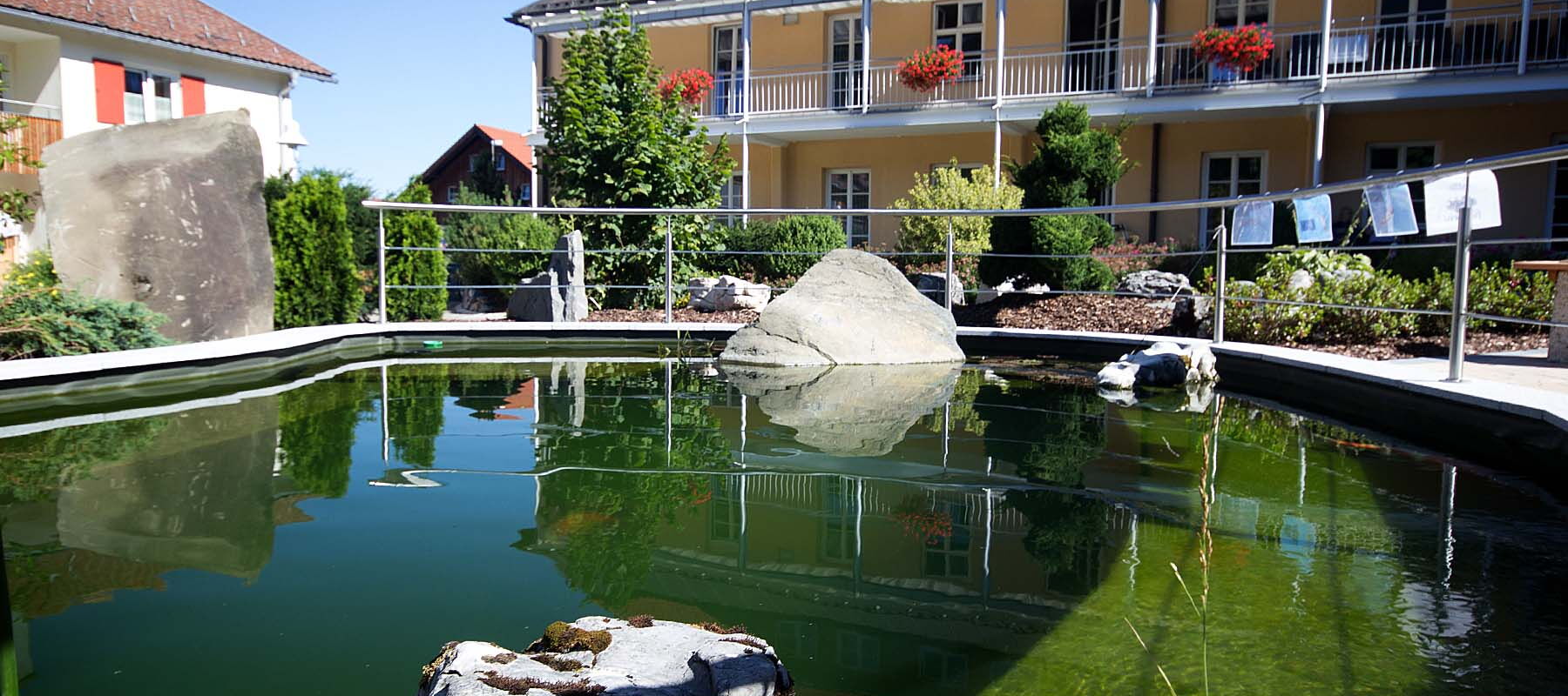 Teich mit großen Steinen im Vordergrund vor einem mehrstöckigen Gebäude mit Balkon und roten Dachziegeln