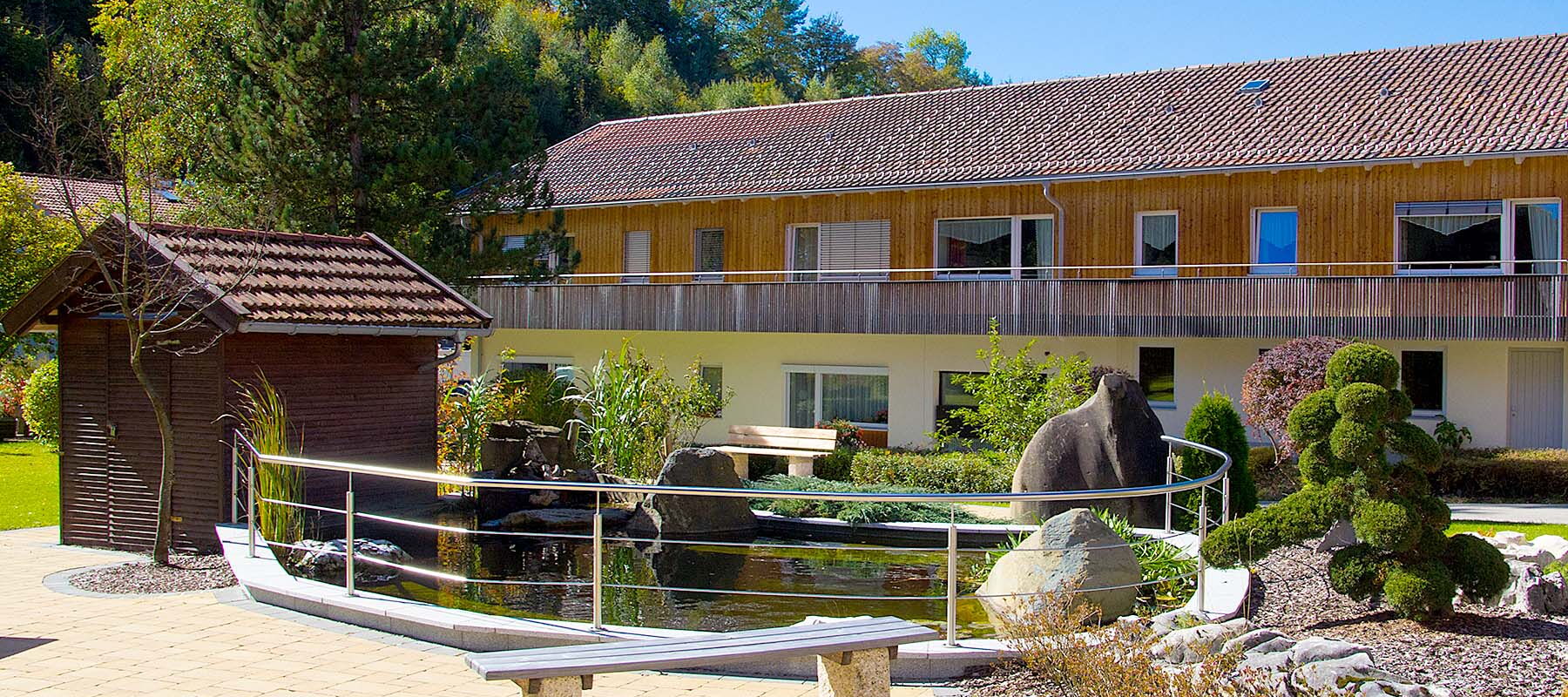 Garten mit Teich, kleiner Holzbrücke, Steinskulptur und zweistöckigem Gebäude im Hintergrund